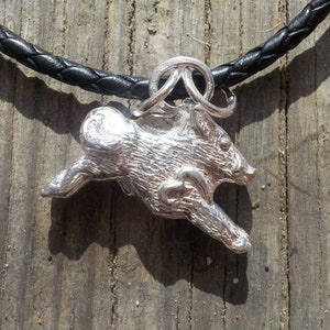 Puede incluir: Un collar con un colgante de plata que representa un jabalí corriendo detallado. El collar está en un cordón de cuero negro.