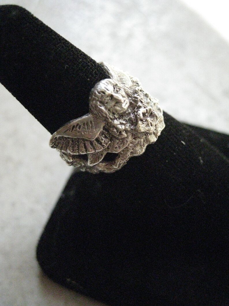 May include: A silver ring with a detailed design of two owls facing each other. The ring is on a black velvet display stand.