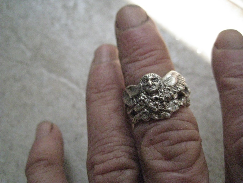 May include: A silver ring with a detailed design of a winged figure with a face. The ring is worn on a finger.