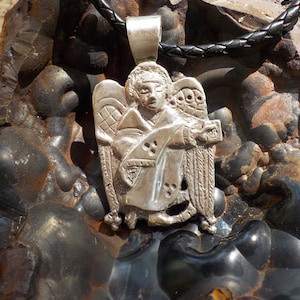 May include: A silver pendant necklace featuring a detailed angel design with wings and a halo. The pendant is attached to a black leather cord.