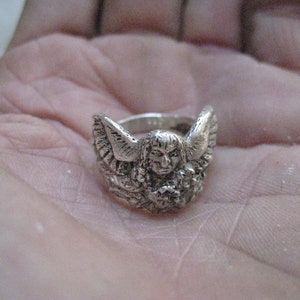 May include: A silver ring with an angel design. The angel has wings and is holding a flower.