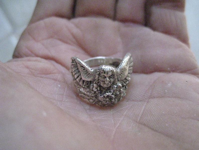 May include: A silver ring with an angel design. The angel has wings and is holding a flower.