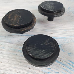 May include: Three black round cabinet knobs with a distressed finish. The knobs have a raised centre and a slightly smaller outer ring.