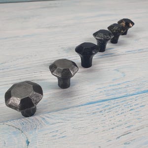 Könnte beinhalten: Eine Sammlung von fünf dunkelgrauen, facettierten Schrankknöpfen. Die Knöpfe haben ein geometrisches Design und eine strukturierte Oberfläche. Sie sind diagonal angeordnet und zeigen ihre einzigartige Form und ihren Stil. Die Knöpfe sind aus Metall.