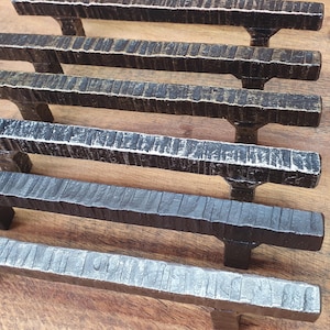 Peut inclure: Une sélection de poignées de meubles en métal noir avec différentes finitions, dont noir et cuivre, noir et or, et noir et argent. Les poignées ont un design rectangulaire texturé. Des étiquettes de texte identifient chaque finition.