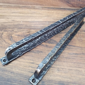 May include: Two rustic, black metal door pulls with a textured, crackled finish. The pulls have a rectangular shape with rounded handles and mounting holes. The metal has a dark, weathered appearance, suggesting a handcrafted or vintage style.