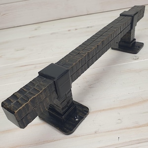 Puede incluir: Un tirador de puerta de metal marrón oscuro y texturizado con un diseño rectangular. El tirador tiene una barra horizontal larga sostenida por dos postes verticales con detalles cuadrados. El acabado tiene un aspecto rústico y envejecido.