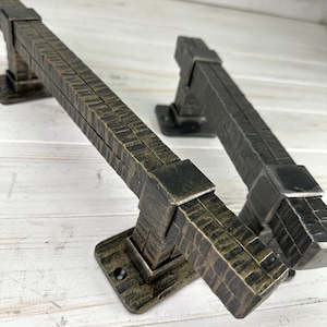 Op de afbeelding: Twee rechthoekige metalen deurklinken met een getextureerde, gehamerde afwerking. De ene klink heeft een donkerbronskleurige patina met gouden accenten, terwijl de andere een donker antracietgrijze afwerking heeft. Beide zijn voorzien van vierkante montageplaten.