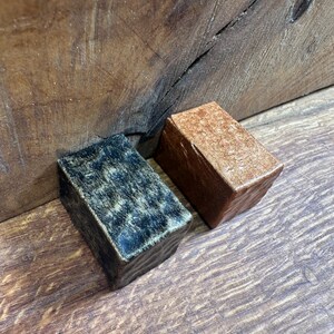 May include: Two rectangular blocks with textured surfaces. One block features a dark, mottled black and gold finish, while the other block has a solid, metallic copper-toned finish. Both items have a slightly uneven, hand-crafted appearance.