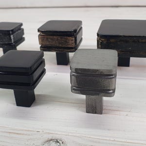 May include: Five different square cabinet knobs with a layered design. The knobs are made of metal and have a variety of finishes, including black, grey, and wood.