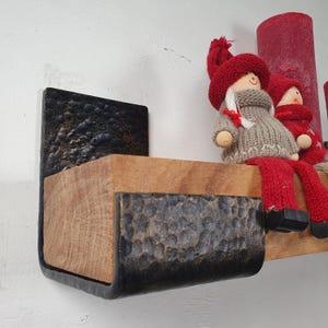 Op de afbeelding: Een wandplank met een houten basis en donkere, getextureerde metalen accenten. De plank bevat twee decoratieve poppen met rode mutsen en truien. Een grote rode kaars draagt bij aan de feestelijke uitstraling.
