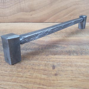 Op de afbeelding: Een lange, rechthoekige, zilverkleurige metalen ladegreep met een gehamerde afwerking. De greep heeft vierkante uiteinden.