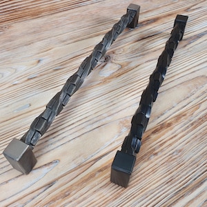 Könnte beinhalten: Zwei schwarze Metall-Schrankgriffe mit einem gedrehten Design. Die Griffe haben quadratische Enden.