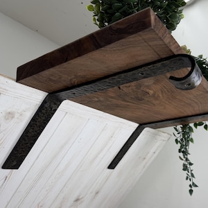May include: A dark brown wooden shelf featuring a natural wood grain texture, supported by a black metal bracket with a hammered, textured finish. The bracket has a curved hook end and is mounted against a white, vertically paneled wall surface.