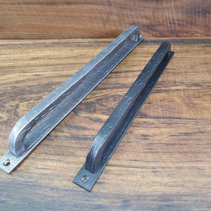 Op de afbeelding: Twee rustieke metalen deurgrepen. Eén is lichtgrijs van kleur, de andere zwart. Beide hebben een getextureerd, gehamerd uiterlijk en een gebogen handvatontwerp. Ze zijn gemonteerd op een houten oppervlak.