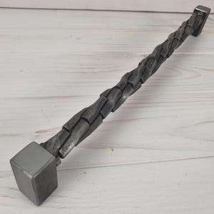 May include: A twisted metal cabinet pull with a square end. The pull is made of dark gray metal and has a textured surface.