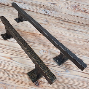 Può includere: Due maniglie per porte in ferro battuto con finitura martellata e testurizzata. Una è bronzo scuro, l'altra è nera. Ognuna ha una barra rettangolare e una piastra di montaggio quadrata. Le maniglie sono su uno sfondo di legno chiaro.