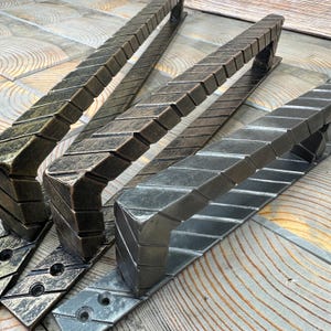 Op de afbeelding: Close-up van verschillende metalen deurgrepen met een getextureerd, rechthoekig ontwerp. De handgrepen hebben een gebogen vorm en zijn in tinten brons en zilver. Ze zijn gerangschikt op een houten oppervlak.