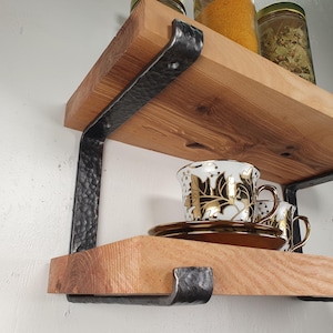 Może przedstawiać: Drewniane półki z czarnymi metalowymi wspornikami. Półki wykonane są z jasnego drewna i wspierane przez młotkowane metalowe wsporniki. Dwie dekoracyjne filiżanki ze złotymi akcentami stoją na dolnej półce. Słoiki z przyprawami są widoczne na górnej półce.