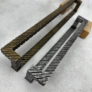 Könnte beinhalten: Zwei rechteckige Türgriffe aus Metall mit einem texturierten, diagonal gerippten Muster. Ein Griff hat ein dunkelbronzenes Finish, während der andere ein silberfarbenes Finish aufweist. Beide Griffe haben eine U-Form mit sichtbaren Montagelöchern.