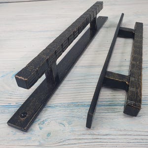 Può includere: Due maniglie per porte in bronzo scuro con un design a blocchi quadrati strutturati. Una è lunga e rettangolare, l'altra è più corta con un design a forma di H. L'hardware è posto su una superficie di legno bianco e azzurro.