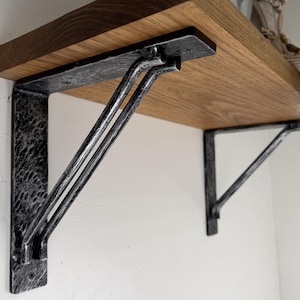 May include: Two L-shaped metal shelf brackets with a hammered, textured black and silver finish supporting a natural light brown wooden shelf. The brackets feature a double-rod diagonal support brace and are mounted to a plain white wall surface.