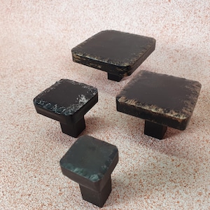 May include: Four square cabinet knobs with a black finish and a distressed look. The knobs are made of metal and have a square base with a rounded top.
