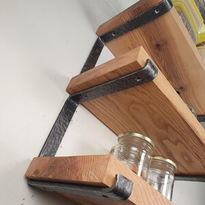 Peut inclure: Étagères en bois avec des supports en métal foncé. Les étagères sont en bois de couleur claire et sont soutenues par des supports métalliques texturés et inclinés. Des bocaux en verre avec des couvercles dorés sont visibles sur les étagères. Les étagères sont fixées à un mur blanc.