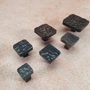 Op de afbeelding: Een set van zes vierkante kastknoppen met een gehamerde metalen afwerking. De knoppen zijn in verschillende tinten zwart en bruin.