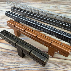 May include: Four rectangular cabinet pulls arranged in a row, each featuring a series of vertical grooves along the top surface. The pulls are finished in distinct colours: light grey, dark charcoal, medium brown, and a dark bronze metallic tone.