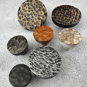 Puede incluir: Una colección de siete pomos circulares para armarios con variados acabados y texturas metálicas. El conjunto incluye piezas con superficies de oro martillado, bronce oscuro, cobre cepillado, plata y negro mate, cada una con patrones únicos.