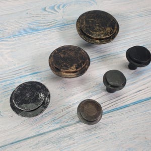 May include: A set of six round cabinet knobs in black and bronze finishes. The knobs have a distressed, aged look and are made of metal.