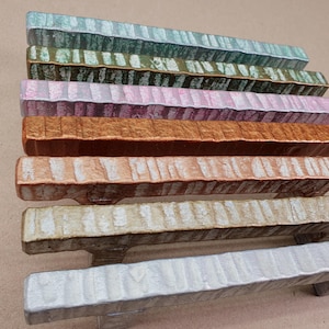 May include: Eight rectangular wooden drawer pulls in a range of colours, including silver, green, pink, and bronze. Each pull has a textured, ridged surface and a small handle underneath. The pulls are arranged in a neat, parallel row, suitable for a chest of drawers.