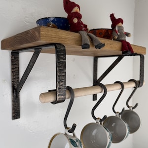 Op de afbeelding: Een wandplank van hout met twee donkere, gehamerde metalen beugels en een horizontale houten stang. Vier zwarte S-vormige metalen haken hangen aan de stang, elk met een keramische kop. Twee kleine decoratieve poppen zitten op het oppervlak van de plank.