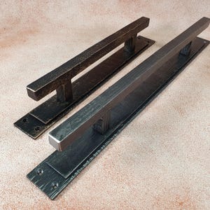 Könnte beinhalten: Zwei schwarze Metall-Türgriffe mit gebürsteter Oberfläche. Die Griffe sind rechteckig mit einem horizontalen Balken in der Mitte. Die Griffe sind auf einer rechteckigen Platte montiert.