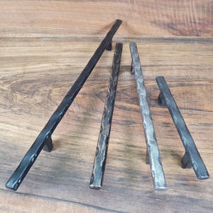 Puede incluir: Cuatro tiradores de armario de metal negro con acabado martillado. Los tiradores son de diferentes longitudes y tienen un aspecto rústico.