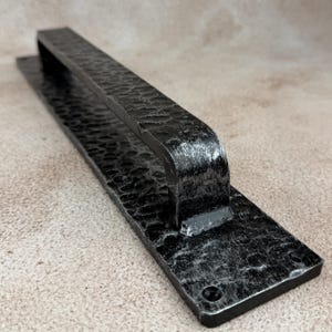 May include: A long, rectangular black metal door handle featuring a hammered, textured surface with silver-toned highlights. The handle is mounted on a flat, matching rectangular backplate with two visible screw holes at each end for installation.