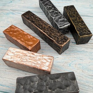 May include: Six rectangular metal bars with varied textured finishes. The collection includes pieces in metallic copper, distressed silver, dark bronze, weathered gold, white-washed copper, and a dark charcoal grey with a hammered dimpled surface.
