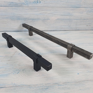 Puede incluir: Dos tiradores para puertas de armario, uno negro y otro gris, con forma rectangular y diseño cuadrado. Los tiradores son de metal y tienen un acabado texturizado.