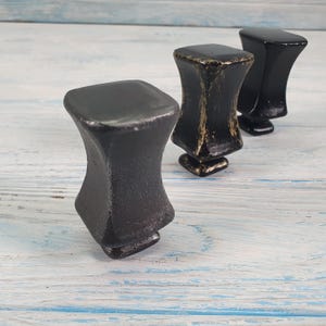 Może przedstawiać: Trzy czarne metalowe gałki do szafek z kwadratowym wierzchołkiem i zaokrąglonym dnem. Gałki mają postarzaną powierzchnię.