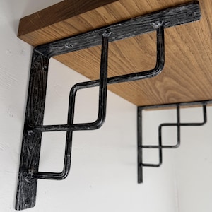 May include: A pair of L-shaped shelf brackets made of dark, textured metal with a distressed finish. Each bracket features a stepped, geometric support design and is attached to a light brown wooden shelf with visible screws at the mounting points.