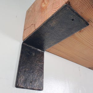 Peut inclure: Un support métallique noir en forme de L avec une surface texturée est fixé à une poutre en bois brun clair. Le support est fixé avec des vis et est conçu pour supporter une étagère ou une autre structure. Le grain du bois est visible.