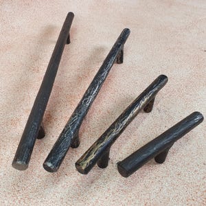 Può includere: Quattro maniglie per armadi in metallo nero con finitura rustica. Le maniglie sono di dimensioni diverse e hanno una forma leggermente curva.