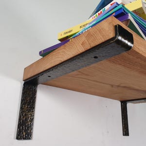 Könnte beinhalten: Ein hellbraunes Holzregal mit schwarzen Metallhalterungen, das eine Reihe von Büchern trägt. Die Halterungen haben eine gehämmerten Textur und sind an einer weißen Wand befestigt.