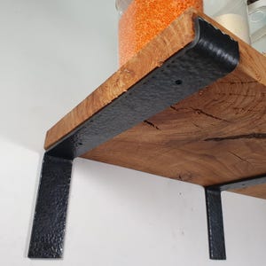 Könnte beinhalten: Rustikales Holzregal mit natürlicher Maserung, getragen von schwarzen, strukturierten Metallhaltern. Gläser mit orangefarbenen Linsen und anderen Gegenständen sind auf dem Regal zu sehen. Geeignet für Küche oder Speisekammer.