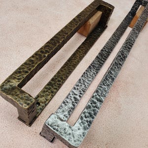 Könnte beinhalten: Zwei Metall-Türgriffe mit gehammerter Textur. Einer ist bronzefarben, der andere silberfarben. Beide sind rechteckig mit einem Griff und auf Holzblöcken montiert. Die Türgriffe sind wahrscheinlich für einen Schrank oder eine Tür.