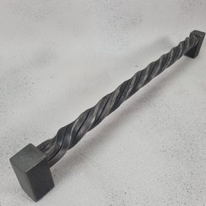 May include: A twisted metal door handle with square ends. The handle is made of dark gray metal and has a rough, textured surface.