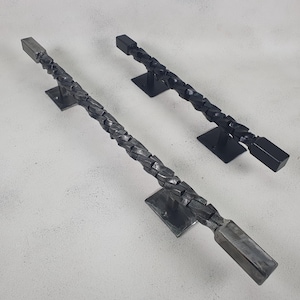 May include: Two black metal door handles with a braided design. The handles are mounted on square plates.