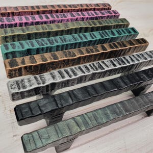 Puede incluir: Una colección de tiradores de armario de estilo rústico en varios colores, incluyendo negro, plata, bronce y verde azulado. Cada tirador tiene un aspecto texturizado y labrado a mano, lo que sugiere una estética artesanal. Los tiradores están dispuestos en fila.