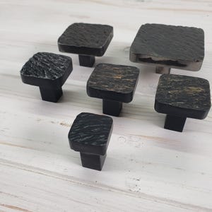 Peut inclure: Six boutons de meubles carrés avec une finition texturée gris foncé. Les boutons ont une base carrée et une courte tige rectangulaire. Les dessus des boutons ont un aspect rugueux et martelé, ajoutant une touche rustique.